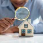 A woman holds a magnifying glass over a miniature model of a house
