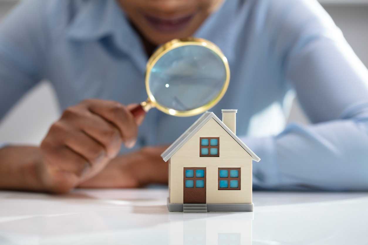 A woman holds a magnifying glass over a miniature model of a house