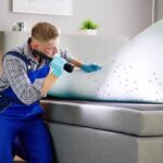 A Bed Bug Exterminator in blue overalls and gloves inspects a mattress with a flashlight, revealing small dark spots on the underside—possible signs of infestation—while focusing on effective solutions for a pest-free home.