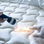 A gloved hand points to small dark spots on a white mattress while another gloved hand shines a flashlight on the area, demonstrating Safe Bed Bug Solutions during a thorough pest inspection.