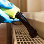 A gloved hand holds a small vacuum cleaner with a brush attachment, providing a fast bed bug solution by cleaning dust from a plastic vent or grid near a window.