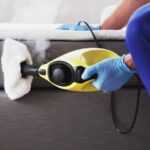 A person wearing blue gloves uses a yellow handheld steam cleaner with a cloth attachment to clean the side of a gray upholstered bed or sofa. Steam is visibly coming from the device.