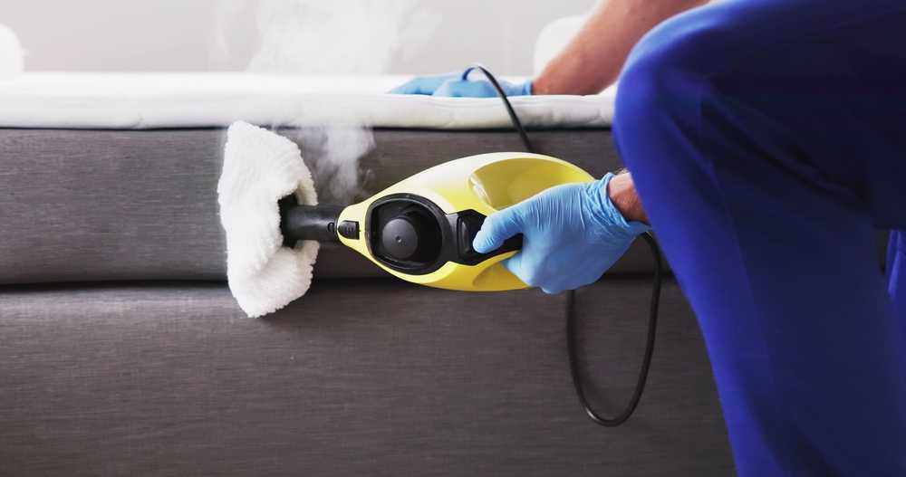 A person wearing blue gloves uses a yellow handheld steam cleaner with a cloth attachment to clean the side of a gray upholstered bed or sofa. Steam is visibly coming from the device.