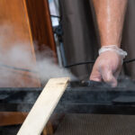 A person wearing a disposable glove uses a steam cleaning device on a black surface, with visible steam rising and a white board propped nearby—an effective bedbug treatment tip for a pest-free home.