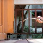 An ant exterminator in an orange shirt uses a spray nozzle to quickly apply pest control treatment at the base of a wooden home structure outdoors.