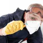 A person wearing a protective suit, yellow gloves, goggles, and a face mask closely examines something with a magnifying glass—an essential step in termite inspections to prevent costly damage. The background is plain and white.