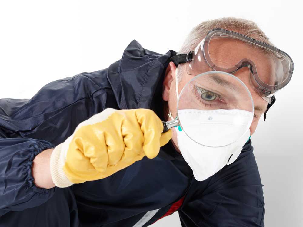 A person wearing a protective suit, yellow gloves, goggles, and a face mask closely examines something with a magnifying glass—an essential step in termite inspections to prevent costly damage. The background is plain and white.