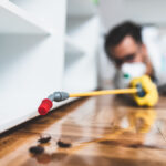 A person wearing yellow gloves uses a spraying tool to apply pesticide near cockroaches on a wooden floor beside a white cabinet, highlighting the work of a cockroach exterminator during indoor pest control activity.