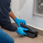 A person wearing blue gloves is placing or inspecting a black rodent bait station on a tiled floor in the corner, demonstrating fast rodent control near a white wall and utility panel.