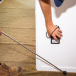 A person in work boots uses a moisture meter on a wall while holding a long probe tool over carpeted flooring next to the wall.