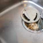 A stainless steel kitchen sink with a drain strainer partially covered by a white lid. Small food scraps are collected in the strainer, which can attract house roaches—keep it clean to help get rid of roaches. Water is running into the sink.