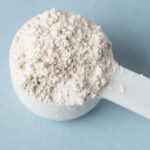 A white plastic scoop filled with fine, white Diatomaceous Earth powder rests on a light blue surface.