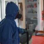 A person in a blue protective suit and mask uses a fogging machine for bed bug fumigation, disinfecting a room with wooden cabinets, a red tablecloth, and an open pantry in the background.