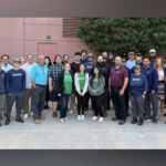 A group of about 35 adults stands outdoors on a paved area in front of a pinkish building and trees, posing for a group photo. Most are casually dressed, and the setting appears to be professional or workplace-related—possibly an event hosted by CERTUS or Termio Pest Control in Arizona.