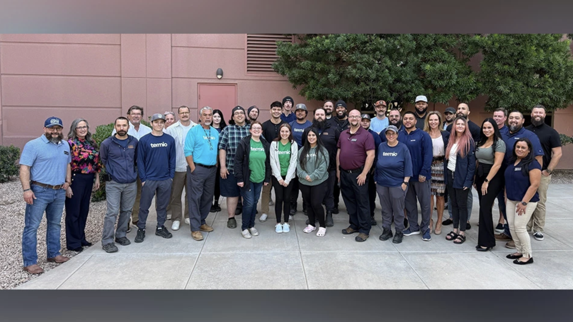 A group of about 35 adults stands outdoors on a paved area in front of a pinkish building and trees, posing for a group photo. Most are casually dressed, and the setting appears to be professional or workplace-related—possibly an event hosted by CERTUS or Termio Pest Control in Arizona.